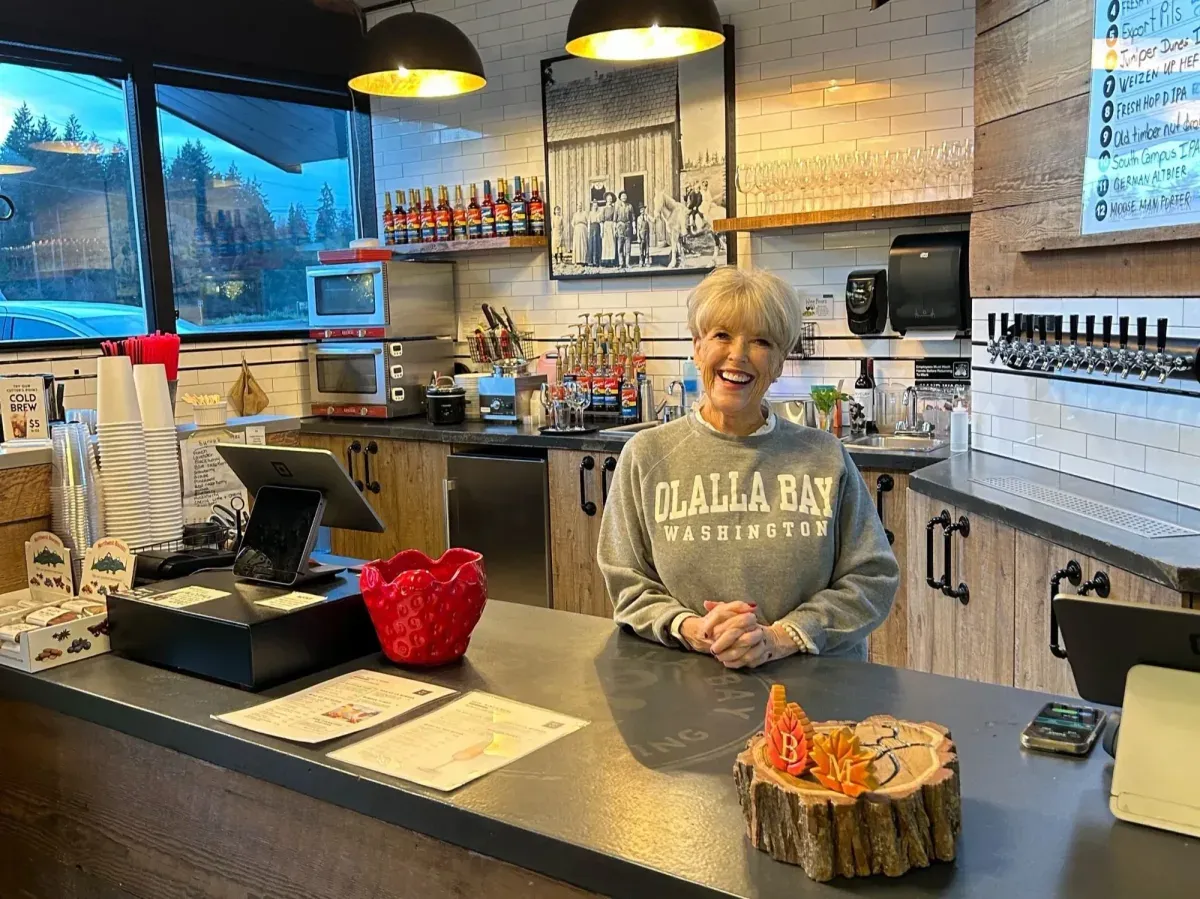A smiling staff member in an Olalla Bay sweatshirt behind the market counter, with the taps and the tap list on the wall