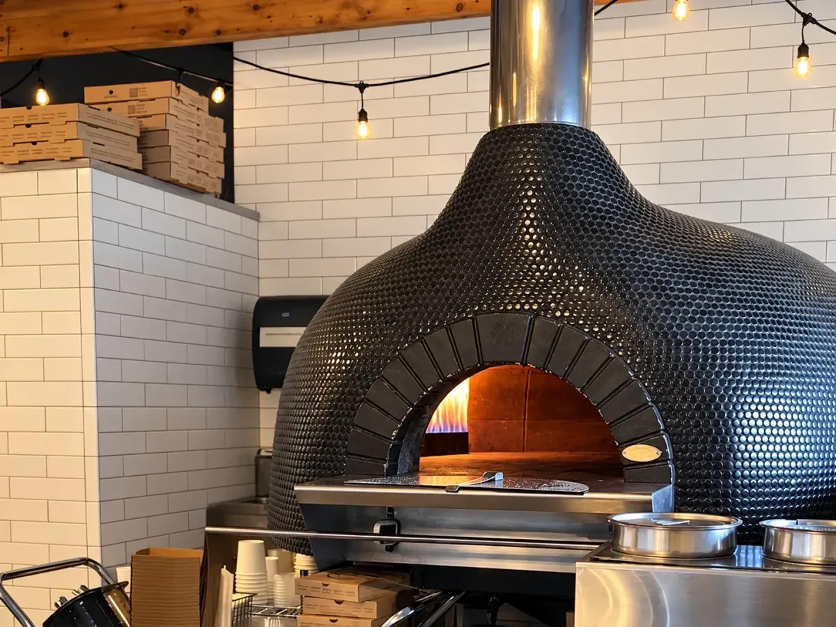 The tiled dome of the pizza oven with a fire glowing inside, under string lights and white subway tile